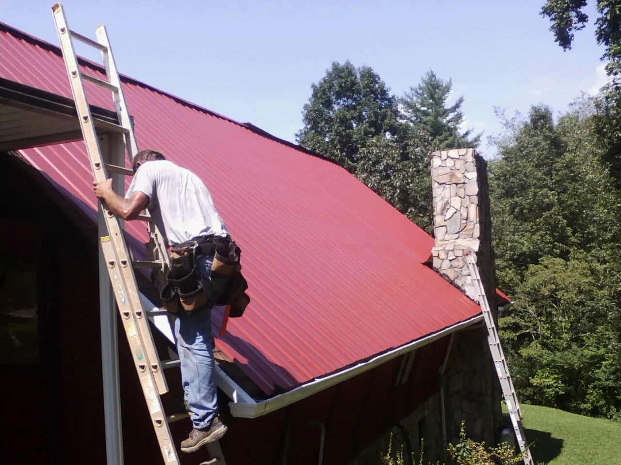 Roofer on a ladder beside a red metal roof and stone chimney — Soffit & Fascia Repair by Roofing Brothers in East Freetown